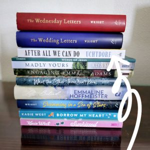 "A neatly stacked pile of books on a dark wooden surface against a light-colored wall. The spines of the books face outward, showcasing their titles and authors. The books include *The Wednesday Letters* and *The Wedding Letters* by Wright, *After All We Can Do* by Uchtdorf, *Words of Jesus* by Condie, *Madly Yours* and *Engaging Emma* by Adams, *What the Other Three Don’t Know* by Hyde, *Left Behind* by Emmaline Hoffmeister, *Swimming in a Sea of Stars* by Wright, *Borrow My Heart* by Kasie West, and *Sunkissed* by Kasie West. A white hand-drawn arrow points to the book *After All We Can Do* by Uchtdorf, emphasizing it. The books appear to be a mix of LDS Christian literature, romance, and contemporary fiction."