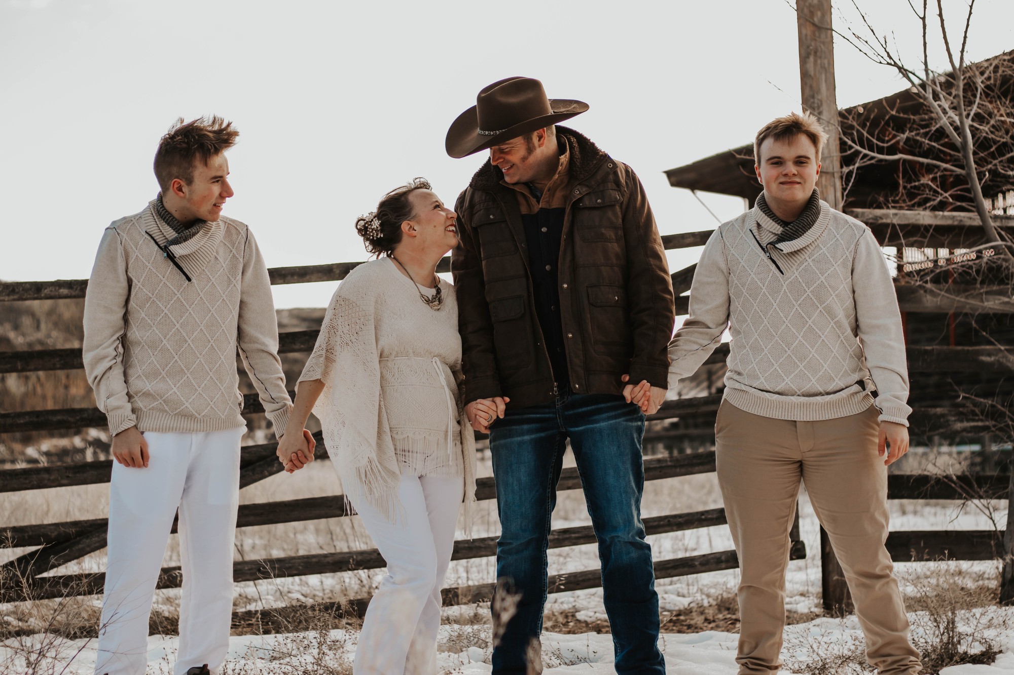 A family portrait outdoors in winter showing two young men, a woman, and a man in a cowboy hat. They are holding hands and smiling, with a rustic wooden fence and barn in the background. The ground is lightly covered in snow, and the family is dressed in coordinated neutral tones with warm sweaters and jackets.