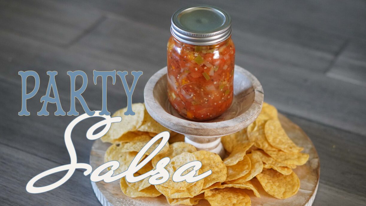 A glass jar filled with homemade party salsa sits on a wooden stand surrounded by crispy tortilla chips, ready for dipping.