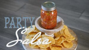 A glass jar filled with homemade party salsa sits on a wooden stand surrounded by crispy tortilla chips, ready for dipping.